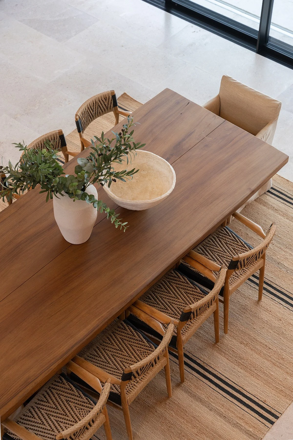 A wooden dining table with a large ceramic vase filled with green foliage and a beige bowl on top. Seven woven chairs are around the table. The room has a light-colored tiled floor, a striped area rug, and floor-to-ceiling glass windows.
