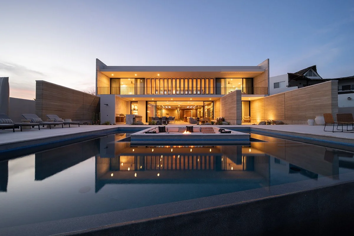 Modern two-story house with large windows, outdoor pool, poolside lounge chairs, and a fire feature at dusk