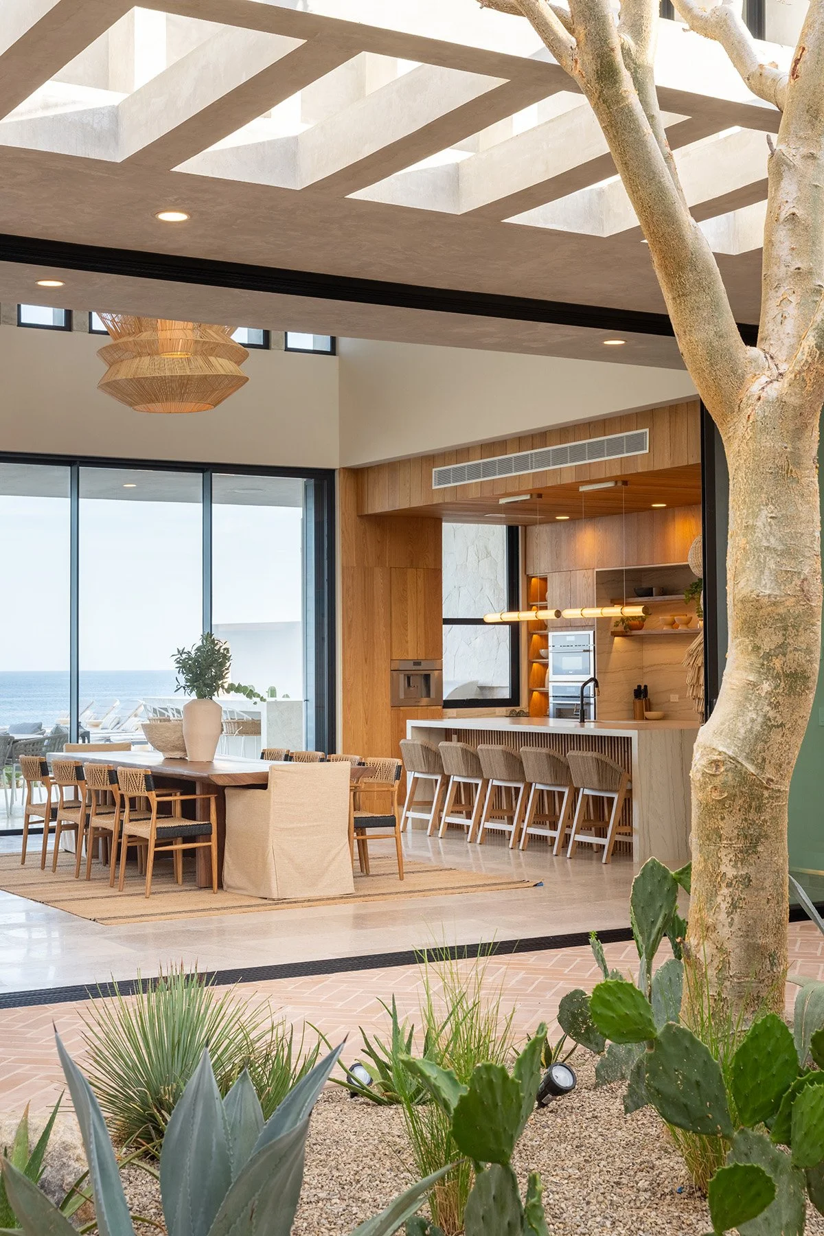 Modern indoor living space with a large dining table, bar counter with seating, large glass windows showing an ocean view, and indoor plants, with natural light and contemporary decor.