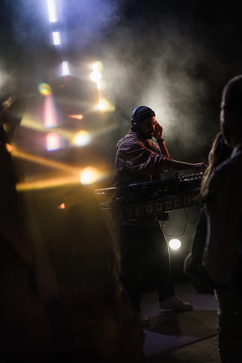 DJ wearing headphones and a cap, mixing music with a DJ controller in a dark club or party setting, with smoke and colorful lights.