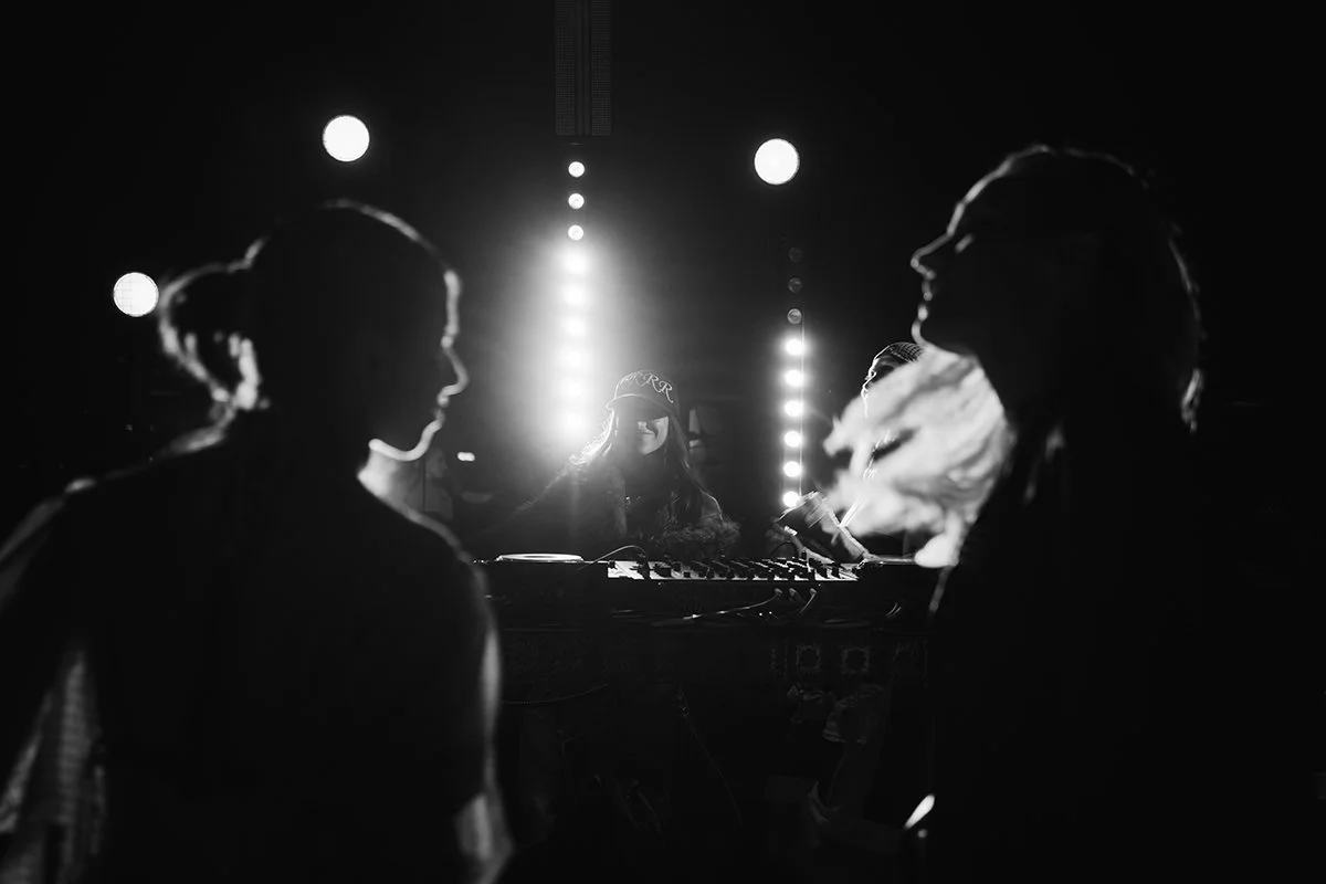 Silhouettes of three women in a dimly lit nightclub, with the DJ at the center illuminated by bright lights behind a DJ booth.