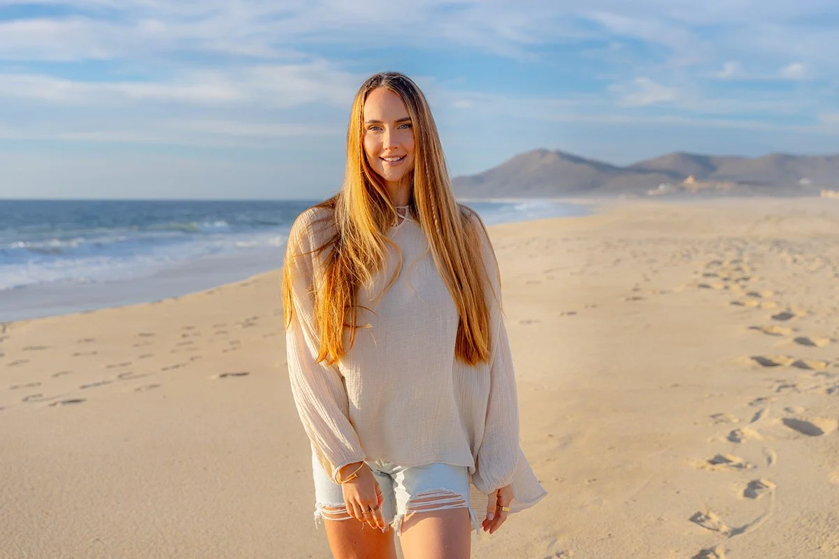 Smiling young woman with long red hair standing on a sandy beach with footprints, ocean waves, and hills in the background during a clear day.