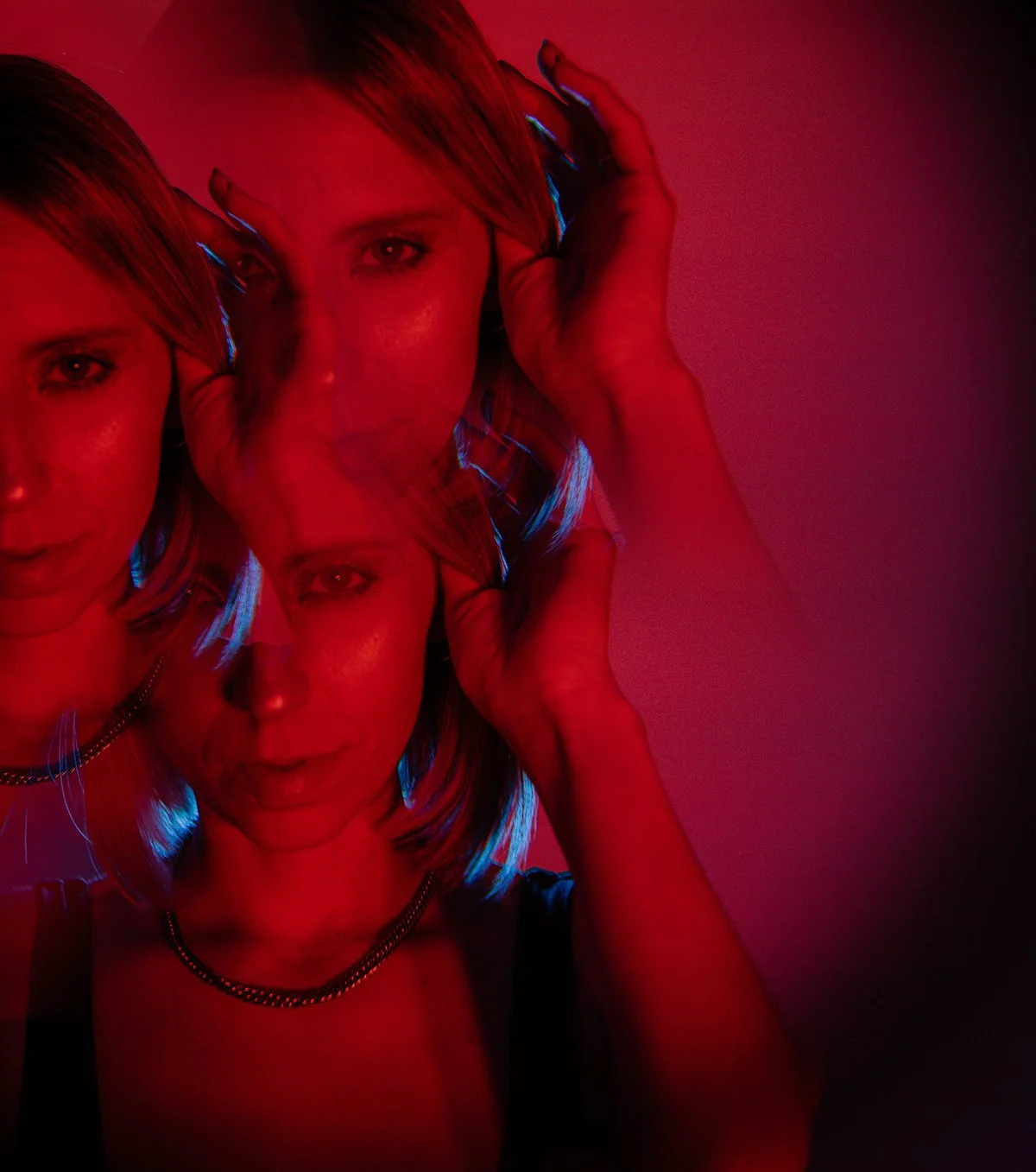 A woman with blonde hair held in her hand, illuminated by red and blue lighting, with a double exposure effect creating overlapping images of her face.