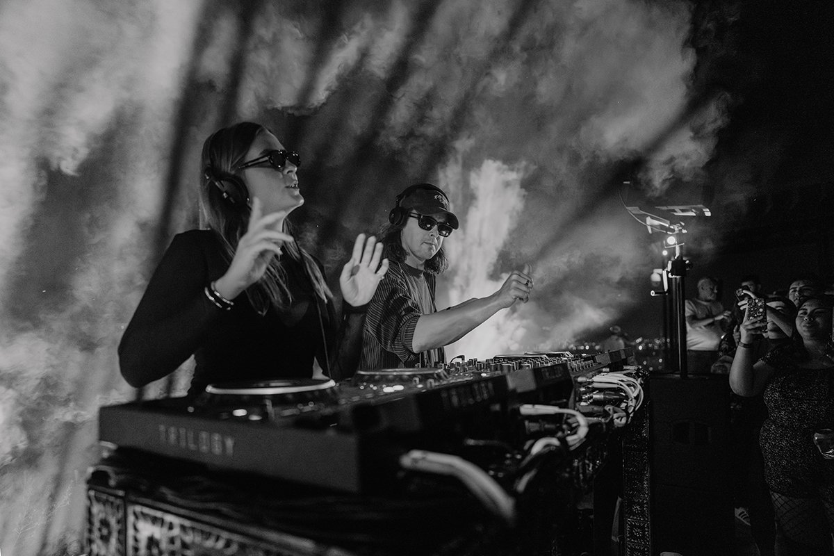 Two female DJs perform at a nightclub, surrounded by a crowd of people, with one wearing sunglasses and headphones, and the other wearing a cap and headphones, against a smoky background.