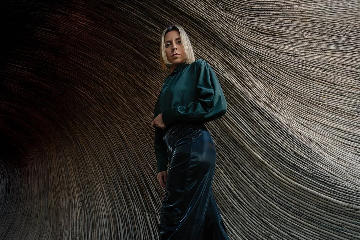 A woman with blonde hair wearing a teal satin bomber jacket and black satin pants stands against a textured, curved wooden wall.
