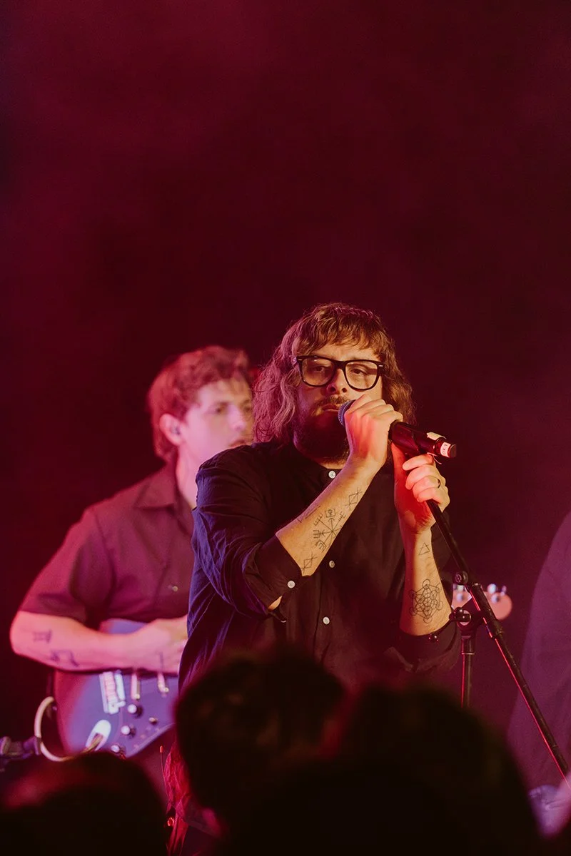 A male singer with glasses and tattoos on his arms holding a microphone during a live performance with red stage lighting. In the background, a guitarist is playing an electric guitar.