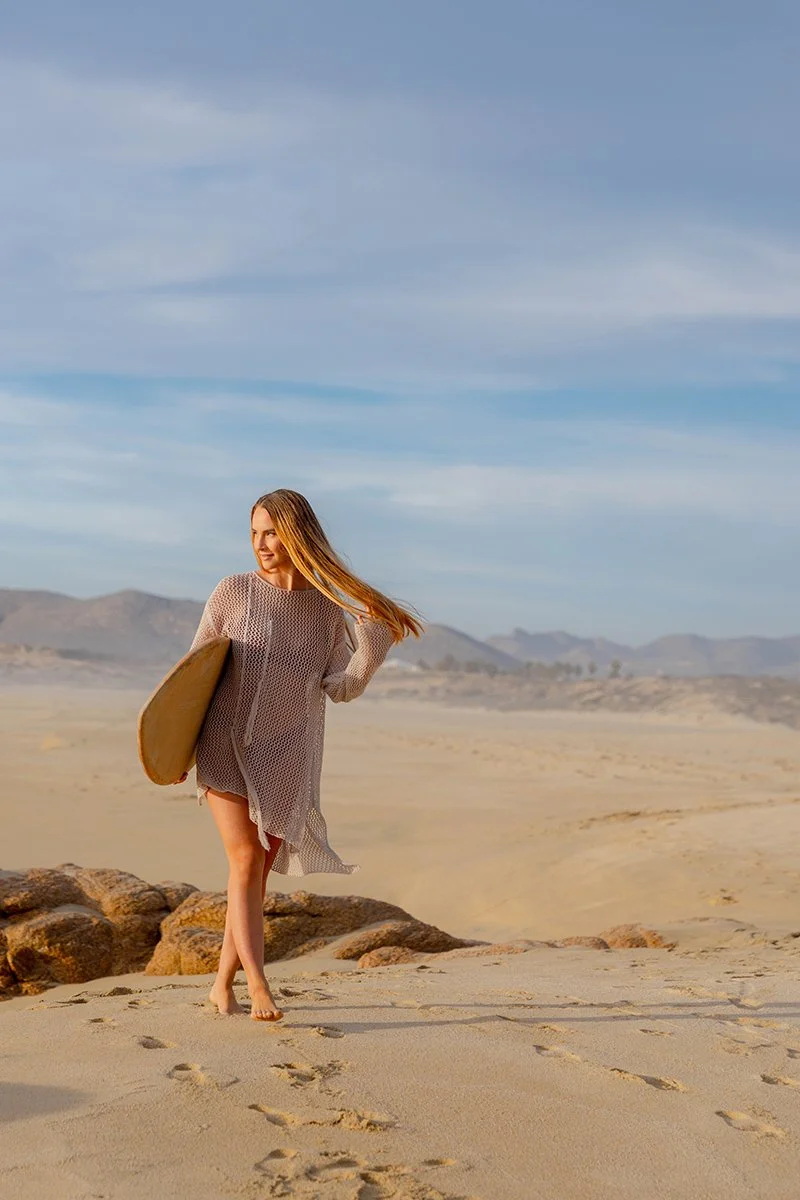 A woman walking barefoot on a sandy beach holding a surfboard, with mountains and a blue sky in the background.