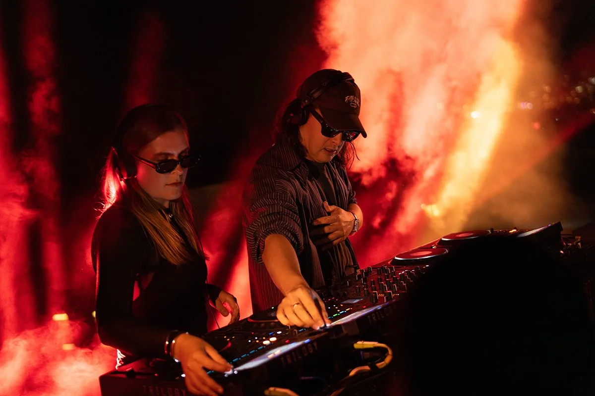 Two DJs performing at a nightclub with red and orange lighting and smoke effects.