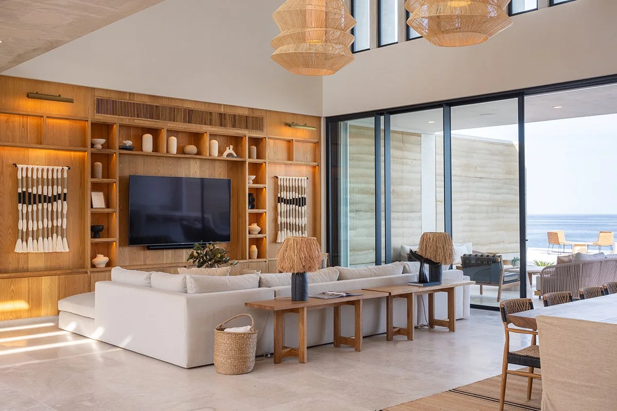 Modern living room with large glass windows, white sectional sofa, wooden coffee tables with lamps, built-in wooden shelves with decor, and ocean view outside.