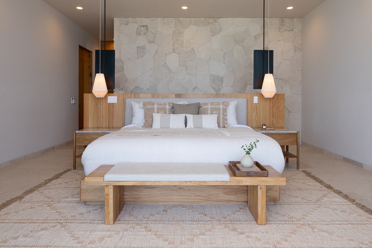 Modern bedroom featuring a large bed with white and beige bedding, decorative pillows, wooden nightstands with hanging pendant lights, a stone accent wall, a wooden bench at the foot of the bed with a small vase and tray, and a beige area rug.