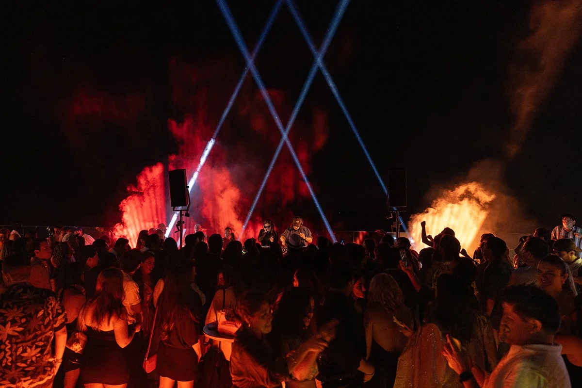 Concert crowd at night with stage lights, smoke effects, and DJ performance, attendees dancing and enjoying music.