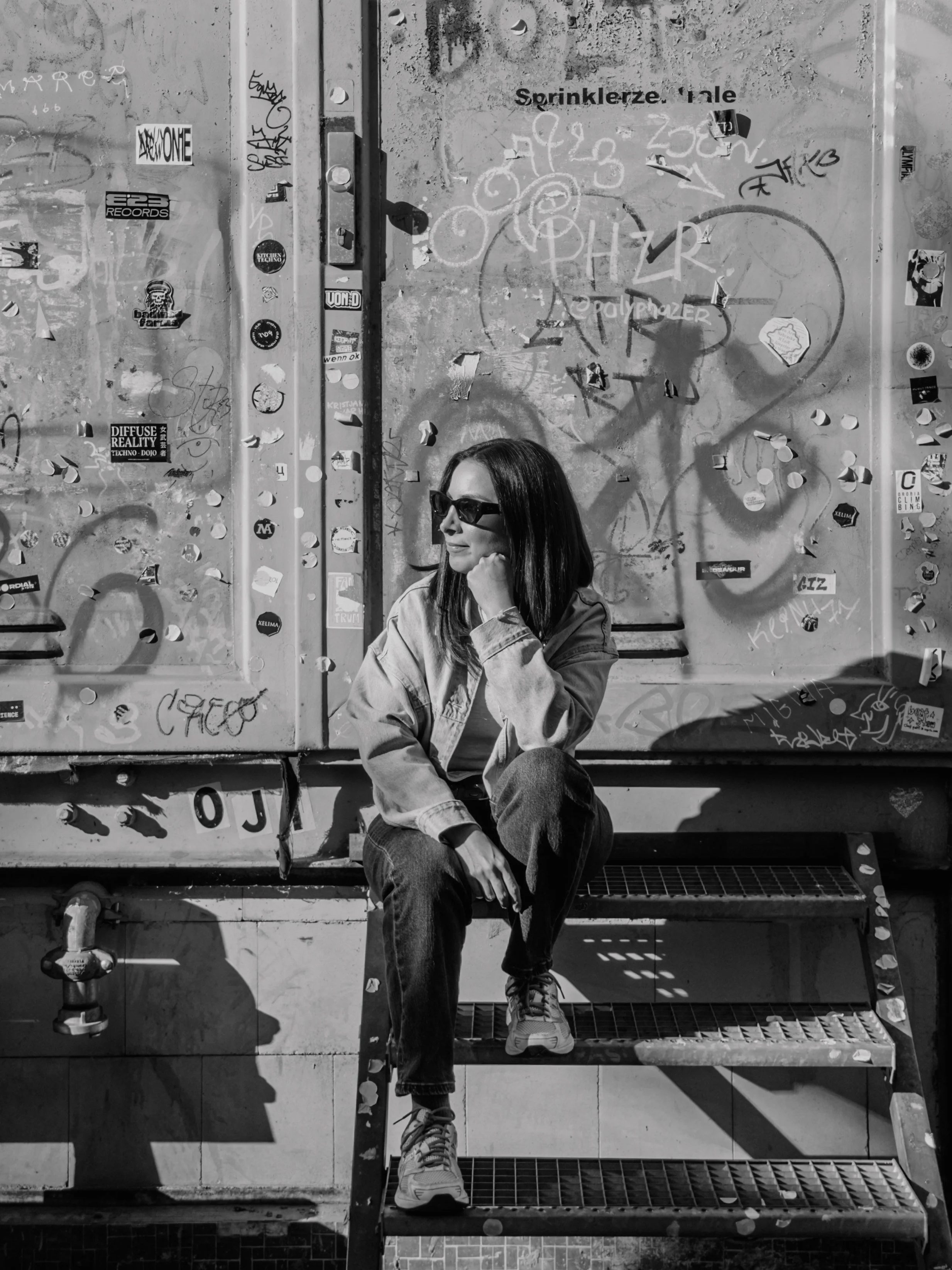 A woman sitting on a metal staircase in front of a graffiti-covered wall. She is wearing sunglasses and a denim jacket, with her head resting on her hand.