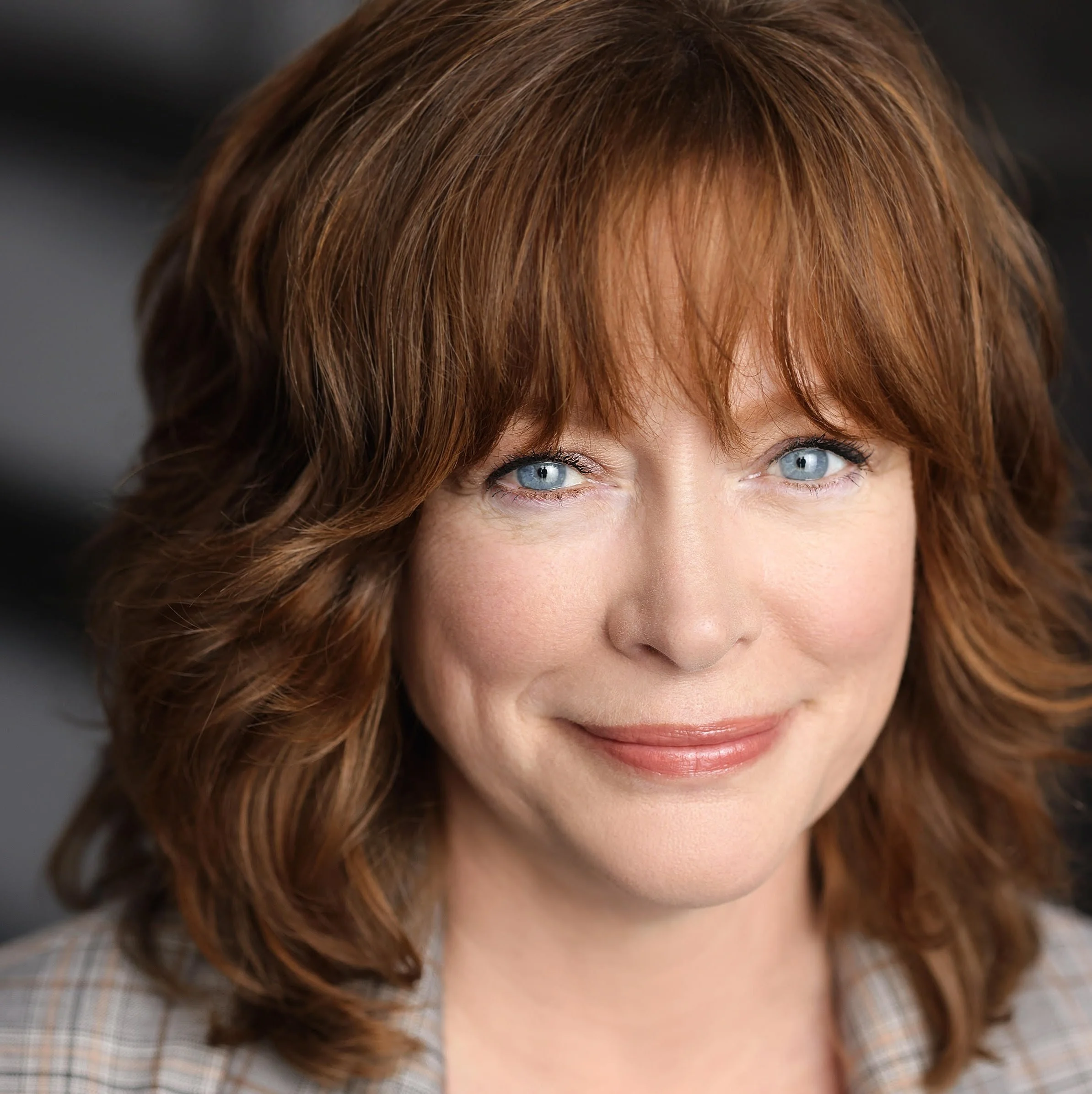 Close-up of a woman with red hair, blue eyes, and light skin, wearing a checked shirt.
