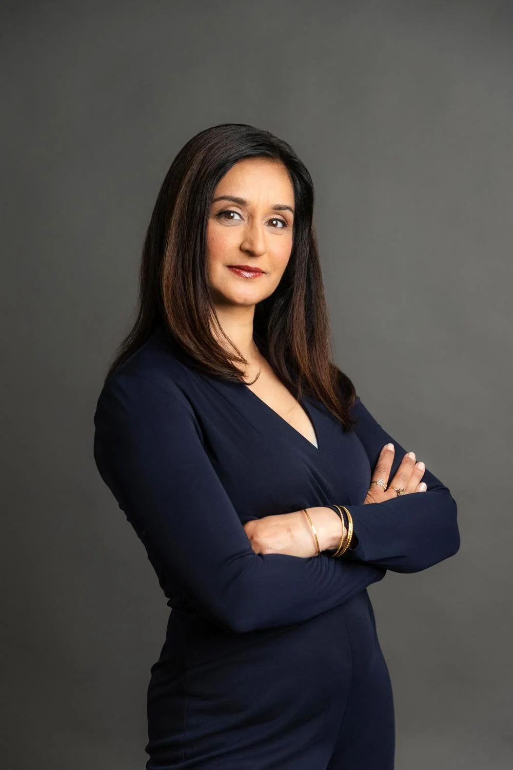 Professional woman with dark brown hair, arms crossed, wearing a navy blue dress and gold bracelets, standing against a gray background.