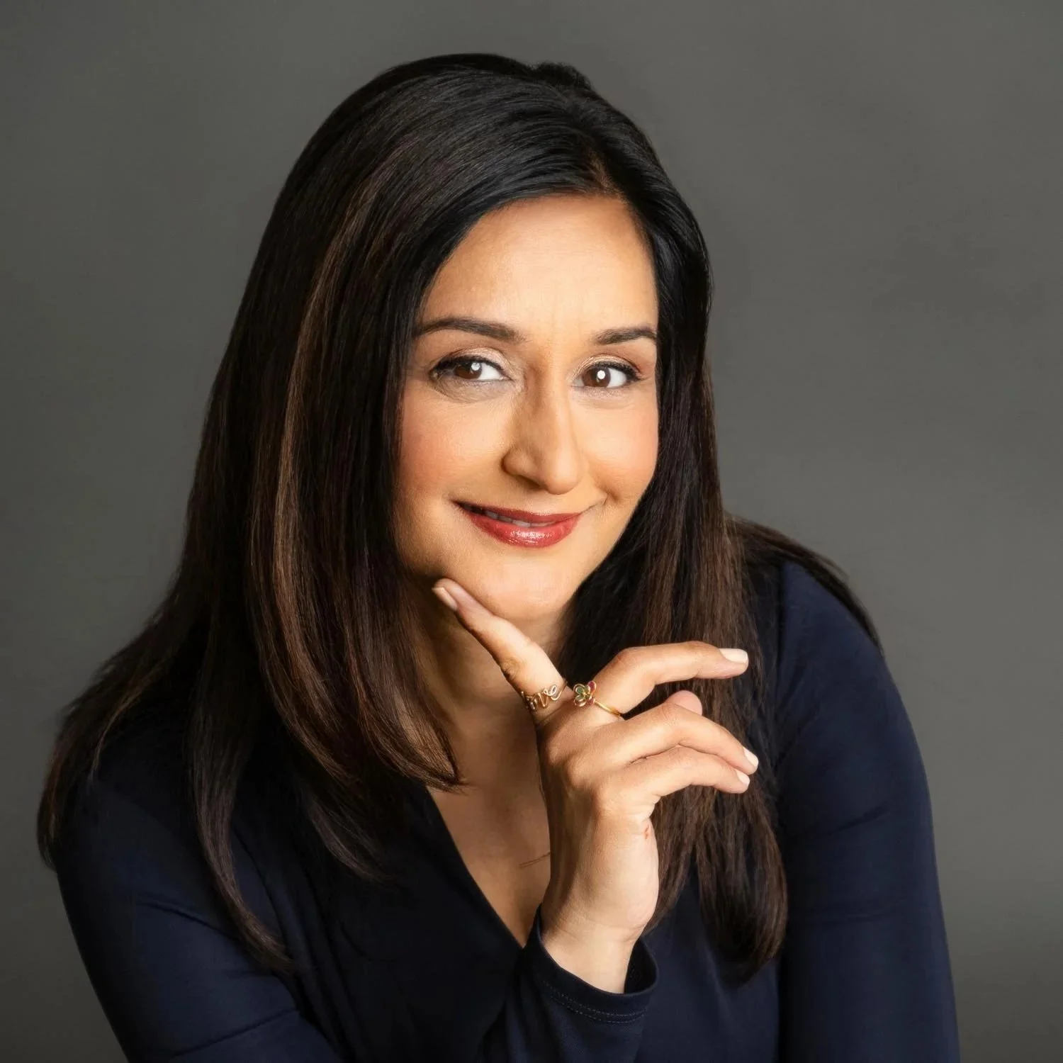 A woman with long dark hair and fair skin, smiling and wearing makeup, is posing with her right hand touching her chin. She is dressed in a dark top with a gray background.