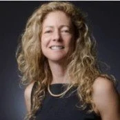 Portrait of a woman with curly blonde hair, wearing a sleeveless black top and a pearl necklace, smiling against a dark background.