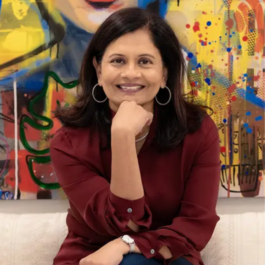 A woman with shoulder-length dark hair, wearing hoop earrings and a maroon blouse, smiling and resting her chin on her hand. She is sitting in front of a colorful abstract mural with splashes of yellow, red, and blue.