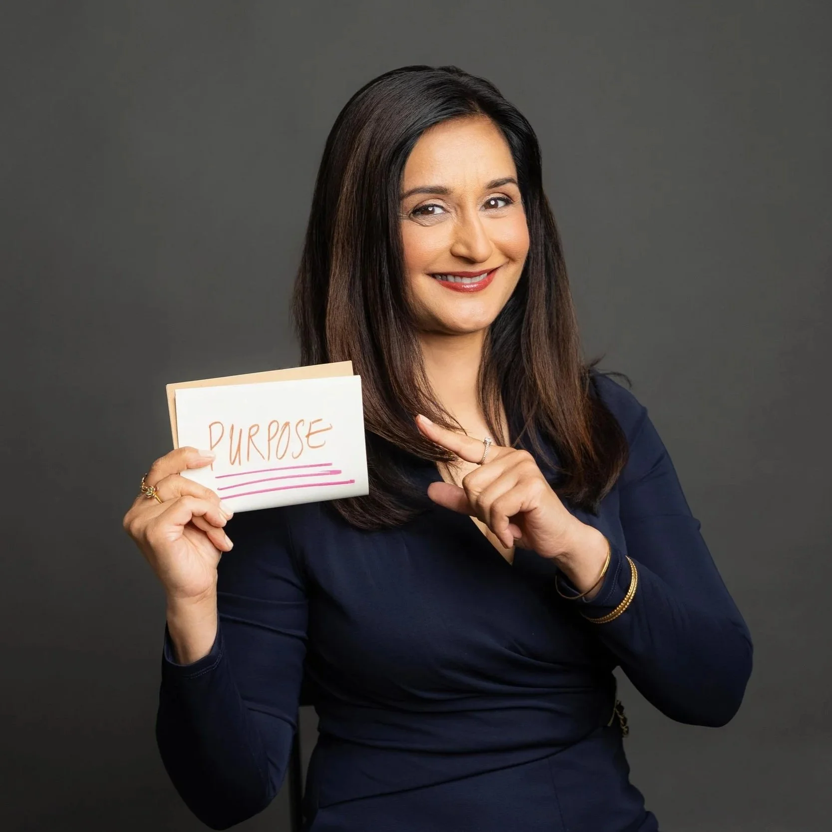 A woman with dark hair smiling and holding a card that says 'Purpose' in front of a gray background.
