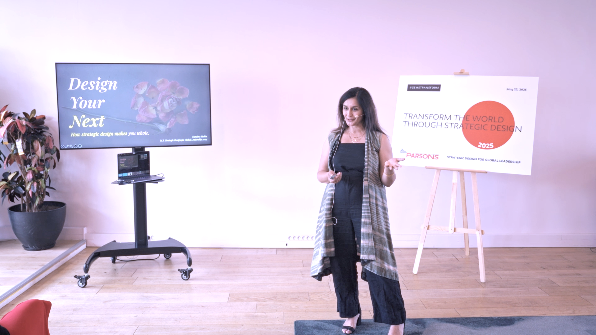A woman giving a presentation, standing near a large screen and a signboard. The screen displays 'Design Your Next' and the signboard reads 'Transform the world through strategic design,' dated May 22, 2025.