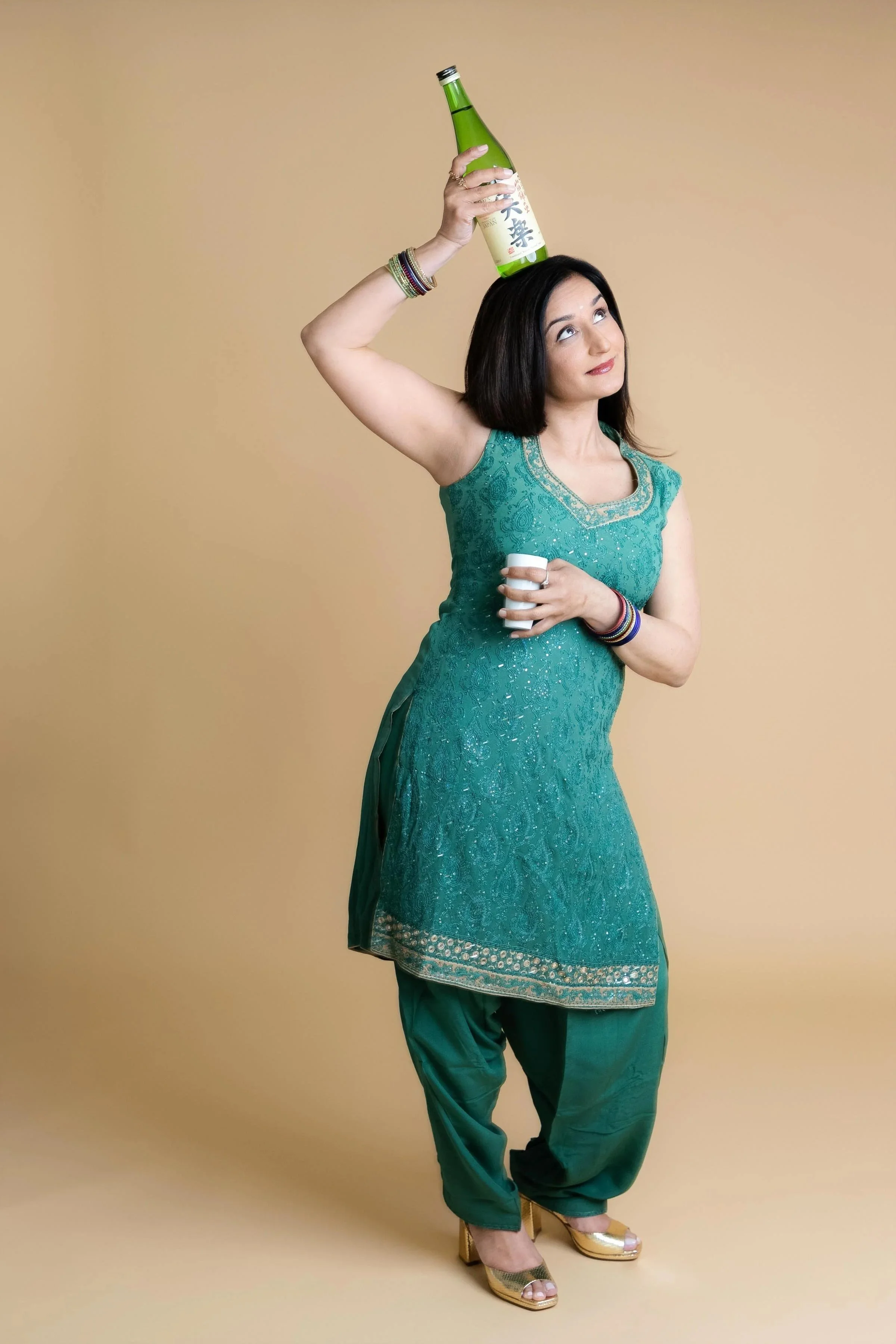 A woman in a green traditional dress holding a beer bottle on her head and a shot glass in her hand, standing against a beige background.