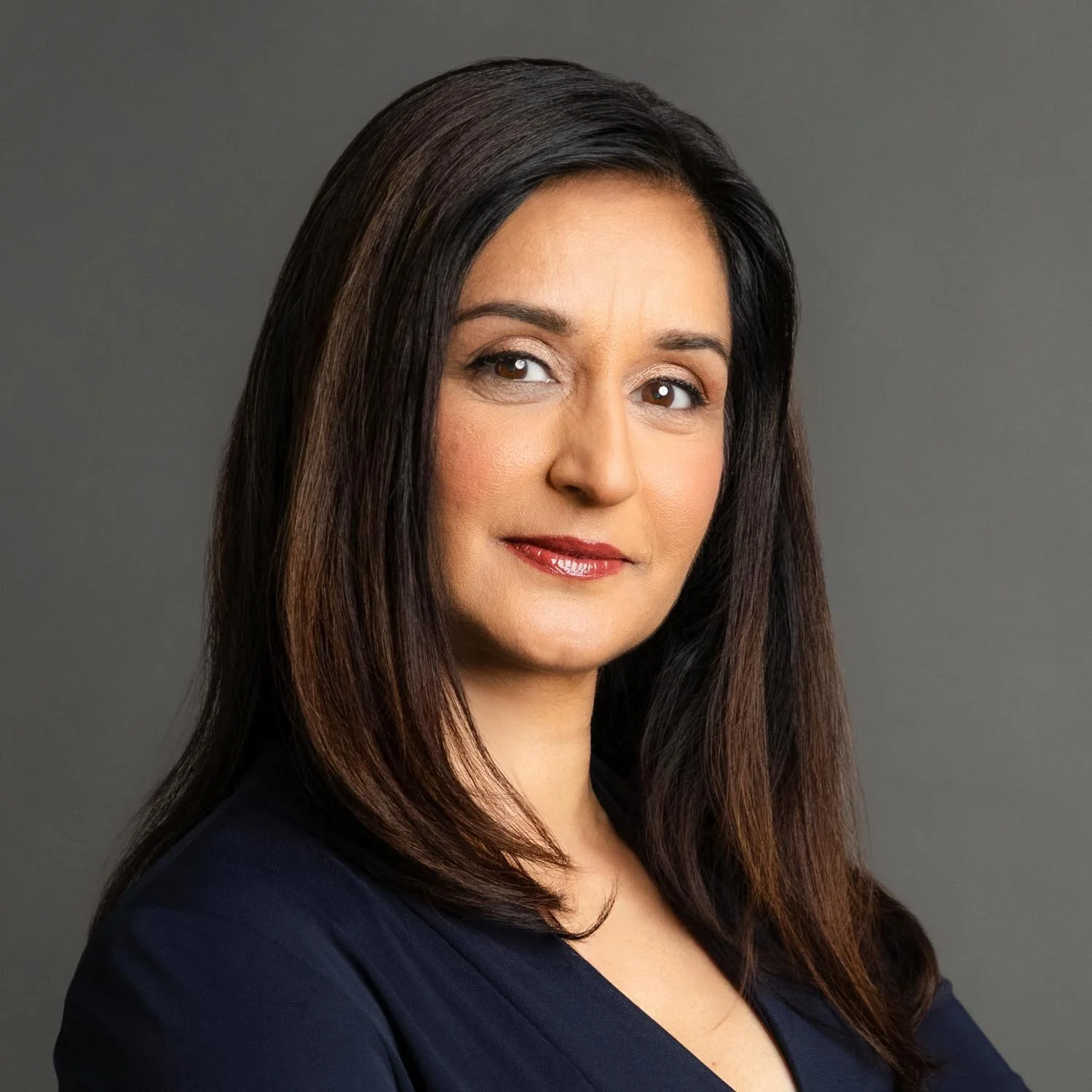 A professional headshot of a woman with long dark brown hair, wearing a dark top, against a gray background.
