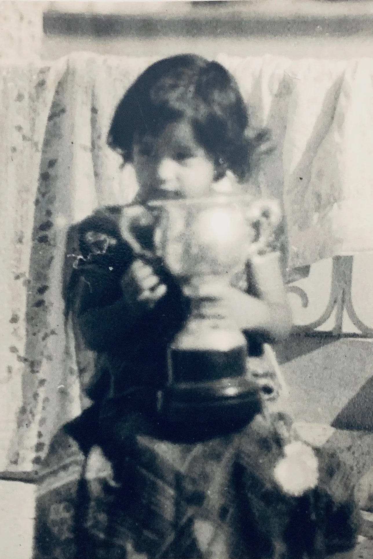 A young child with curly hair taking a selfie in a mirror, holding a camera or phone, sitting on a chair with a floral pattern, with a curtain in the background.