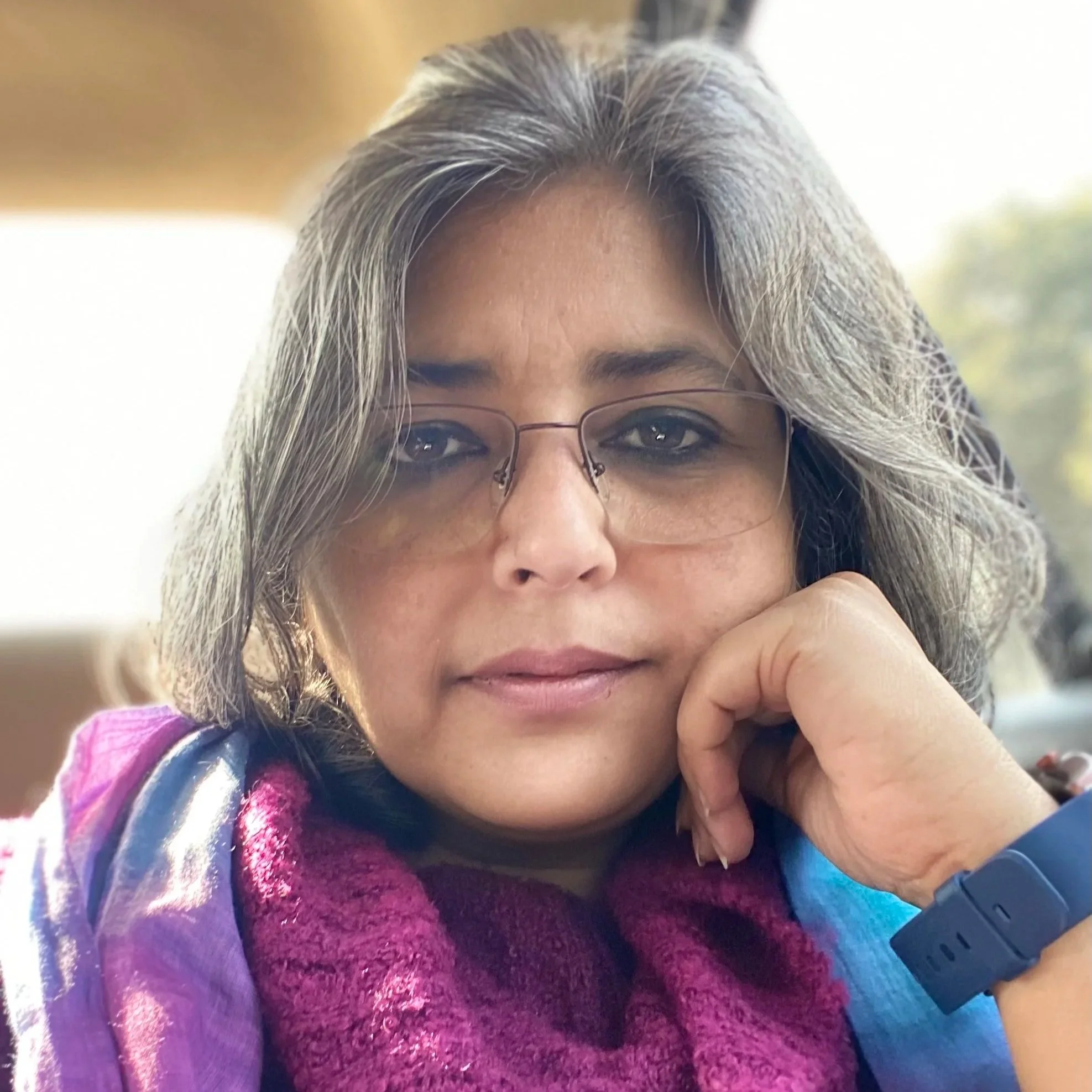 A woman with gray hair and glasses, resting her chin on her hand, wearing a purple and pink knitted scarf, with a blurred background.