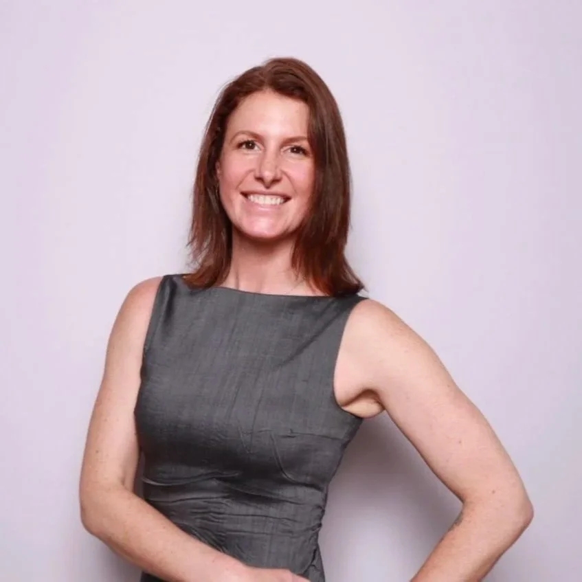A woman with red hair in a sleeveless gray dress, smiling with her right hand on her hip against a plain white background.