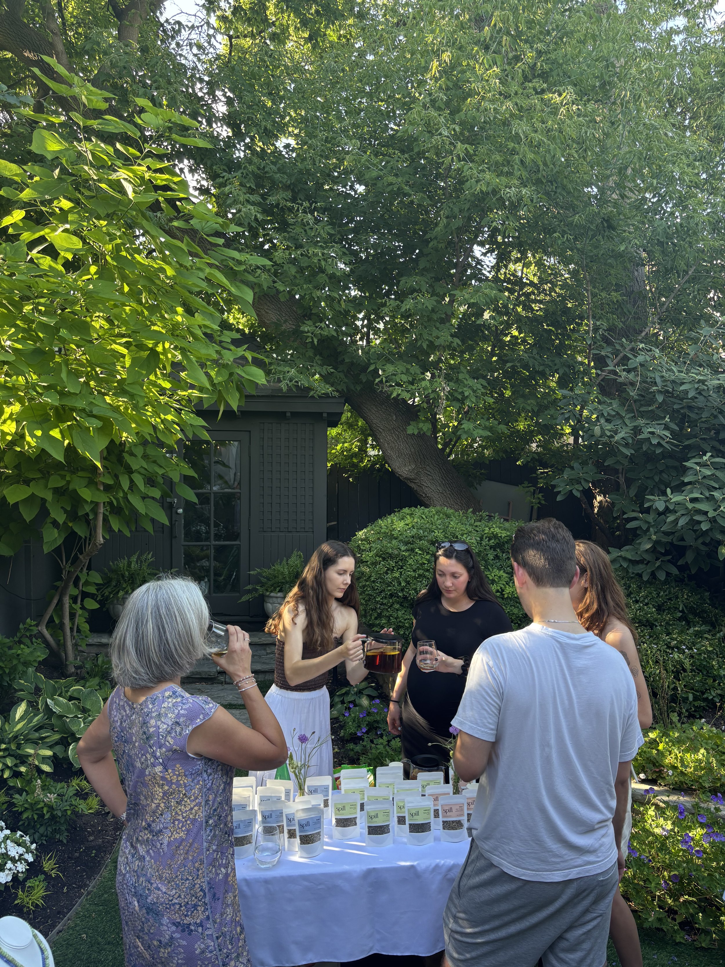 Group of five people at an outdoor gatherign, examining and pouring wine from bottles on a table, with lush green trees and plants in the background.