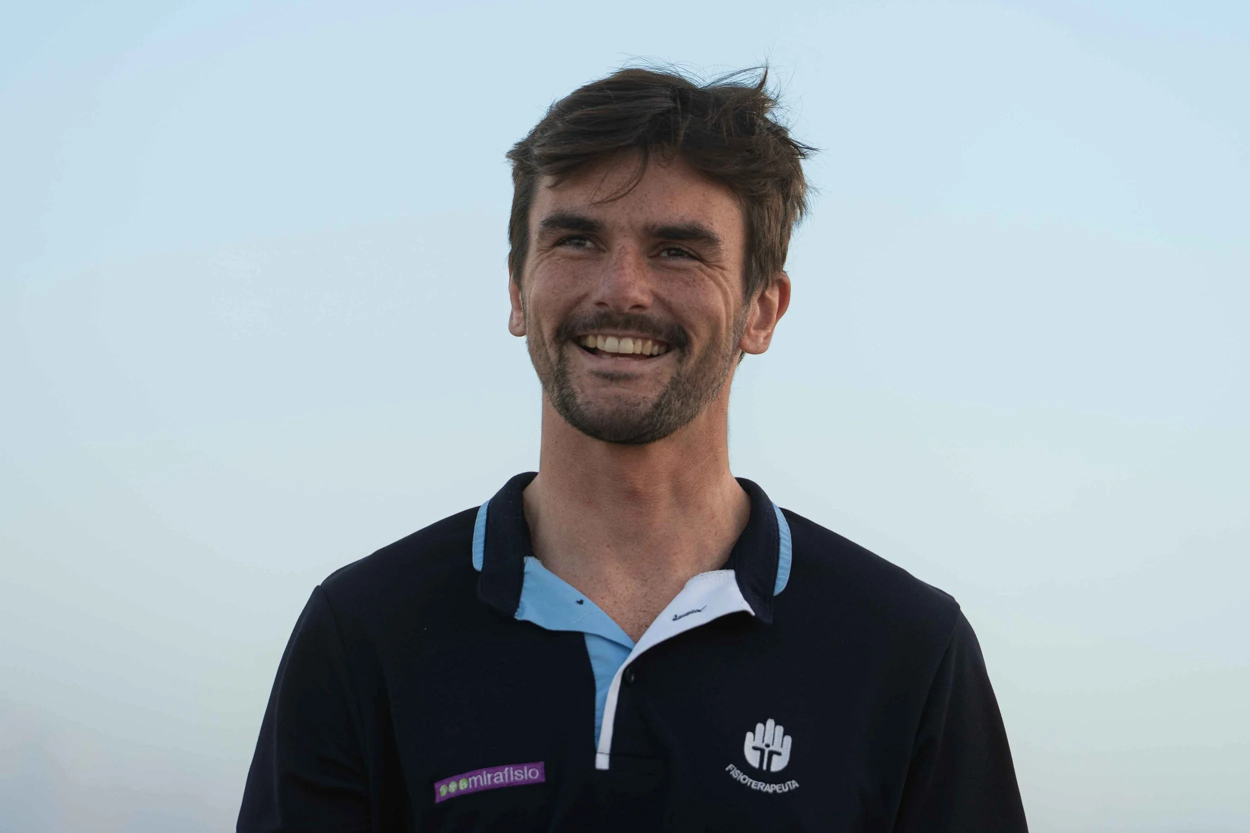 Un homme souriant avec une barbe porte un polo noir avec logo de physiothérapeute, fond de ciel clair.