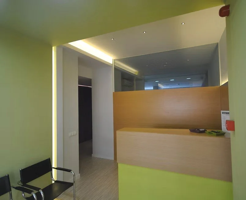 Vue de l'intérieur d'une salle ou d'une réception avec un comptoir en bois, un mur en verre, et deux chaises noires au premier plan.