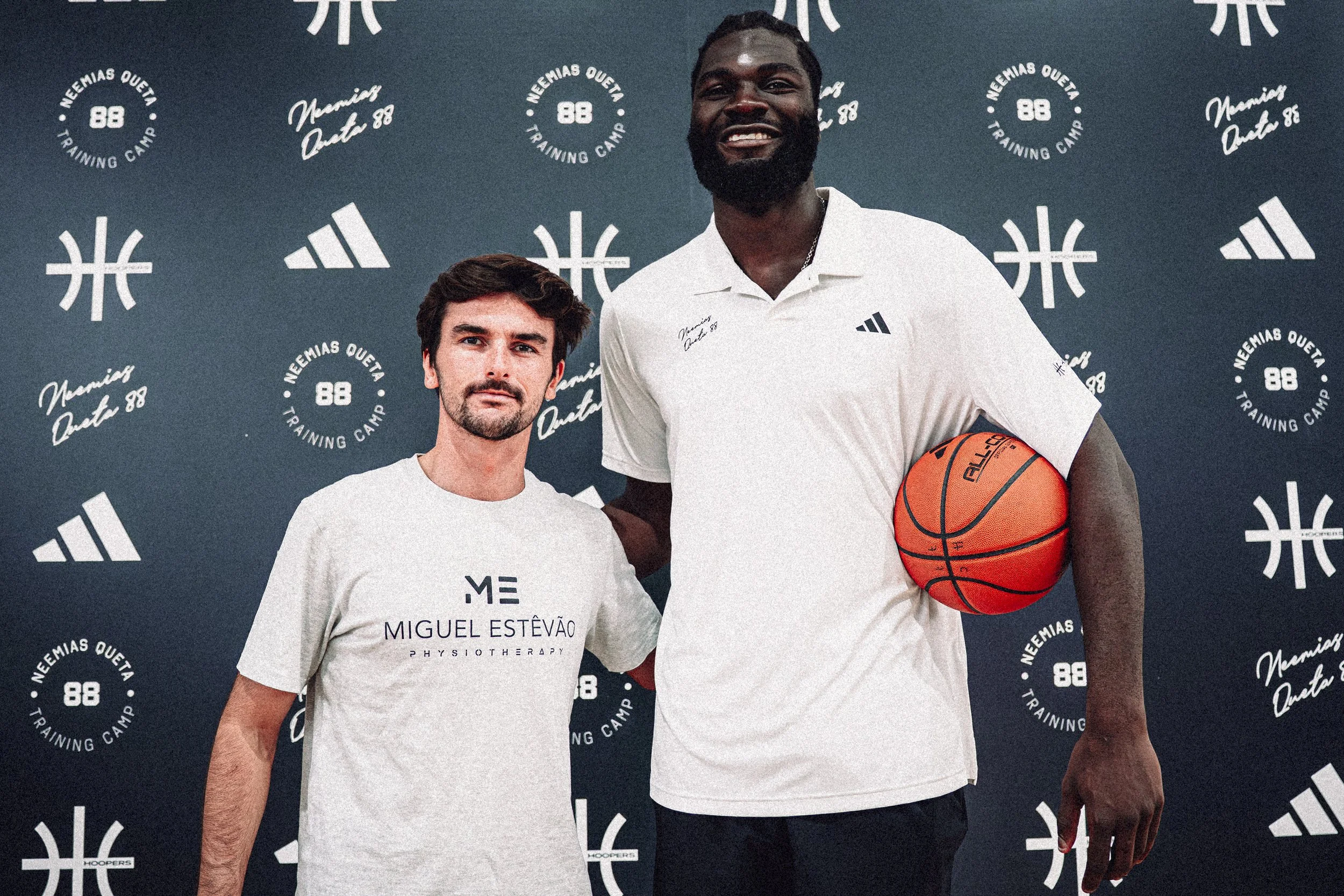 Deux hommes posent devant un fond avec le logo 'Neemias Queta Training Camp 88' et des logos Adidas. L'un porte un t-shirt blanc avec le nom 'Miguel Estêvão' et une mention 'Physiotherapy', l'autre porte un polo blanc avec un logo Adidas et tient un ballon de basketball orange.
