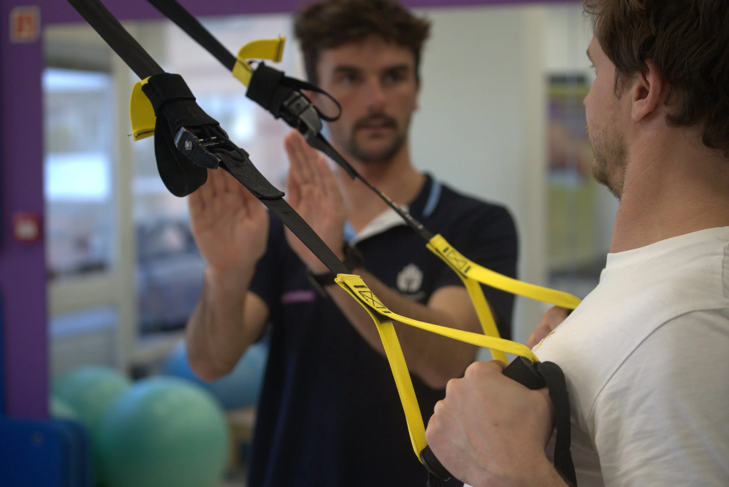 Deux hommes s'entraînent à l'utilisation d'une sangle de soutien ou d'entraînement avec un professionnel dans un centre de rééducation ou de physiothérapie.