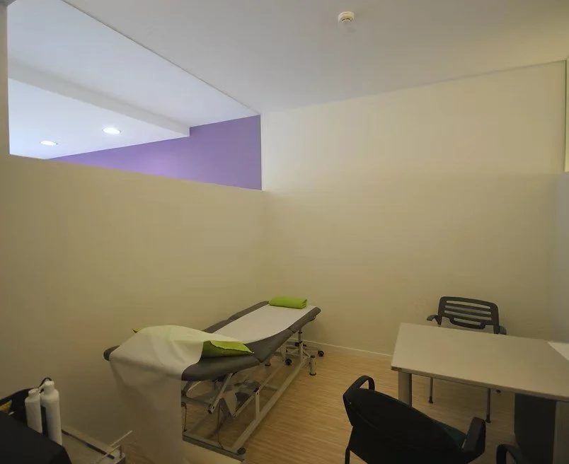 Salle de consultation médicale avec un lit d'examen, un bureau, deux chaises et un mur blanc, avec un plafond en partie ouvert et une lumière encastrée.