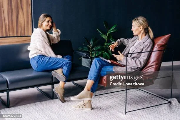 Two women having a conversation in a modern sitting area with a black couch, a plant, and a textured rug.