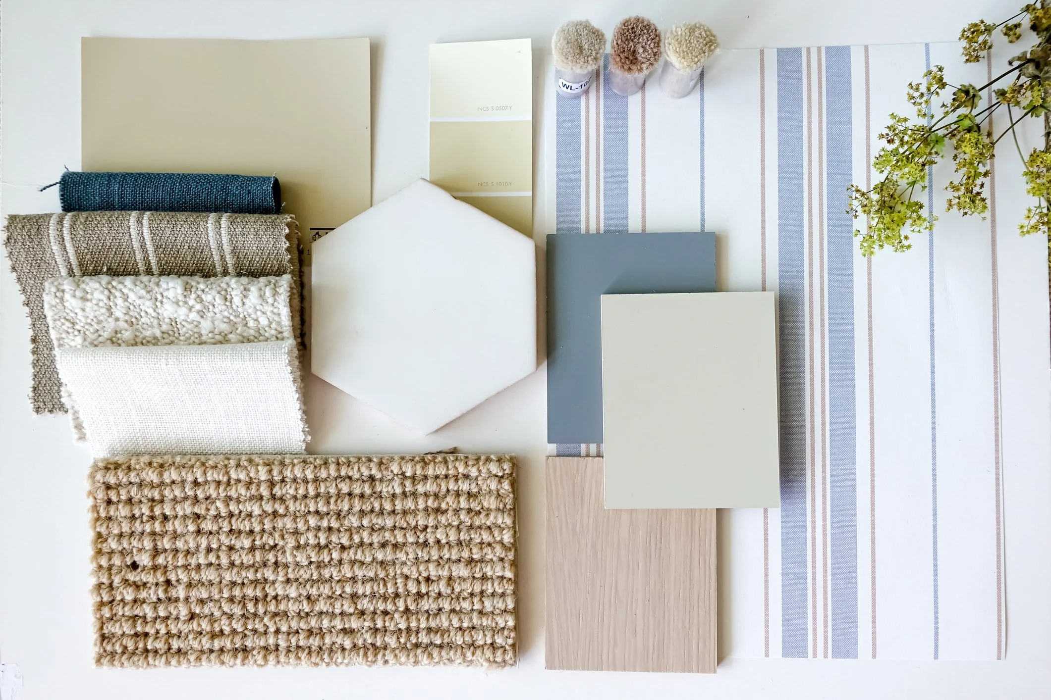 Samples of fabric, paint swatches, paint chips, a hexagon and rectangular wall tiles, a wood plank, a floral patterned tile, small potted plants, and a striped table runner on a white surface.