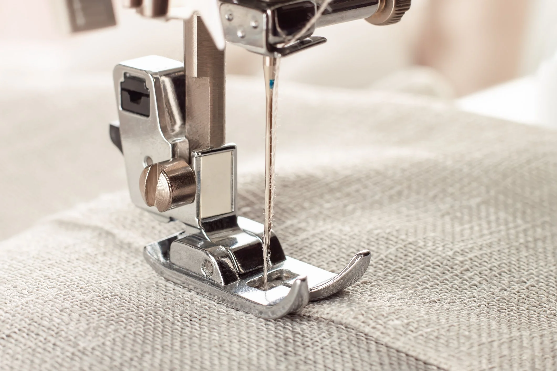 Close-up of a sewing machine needle and presser foot stitching beige fabric.