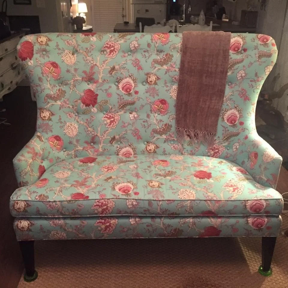 A vintage-style sofa with a floral pattern in pink, red, and beige on a light blue background. A pinkish-brown throw blanket is draped over the back of the sofa. The sofa is situated in a living room with warm lighting and other furniture visible in the background.
