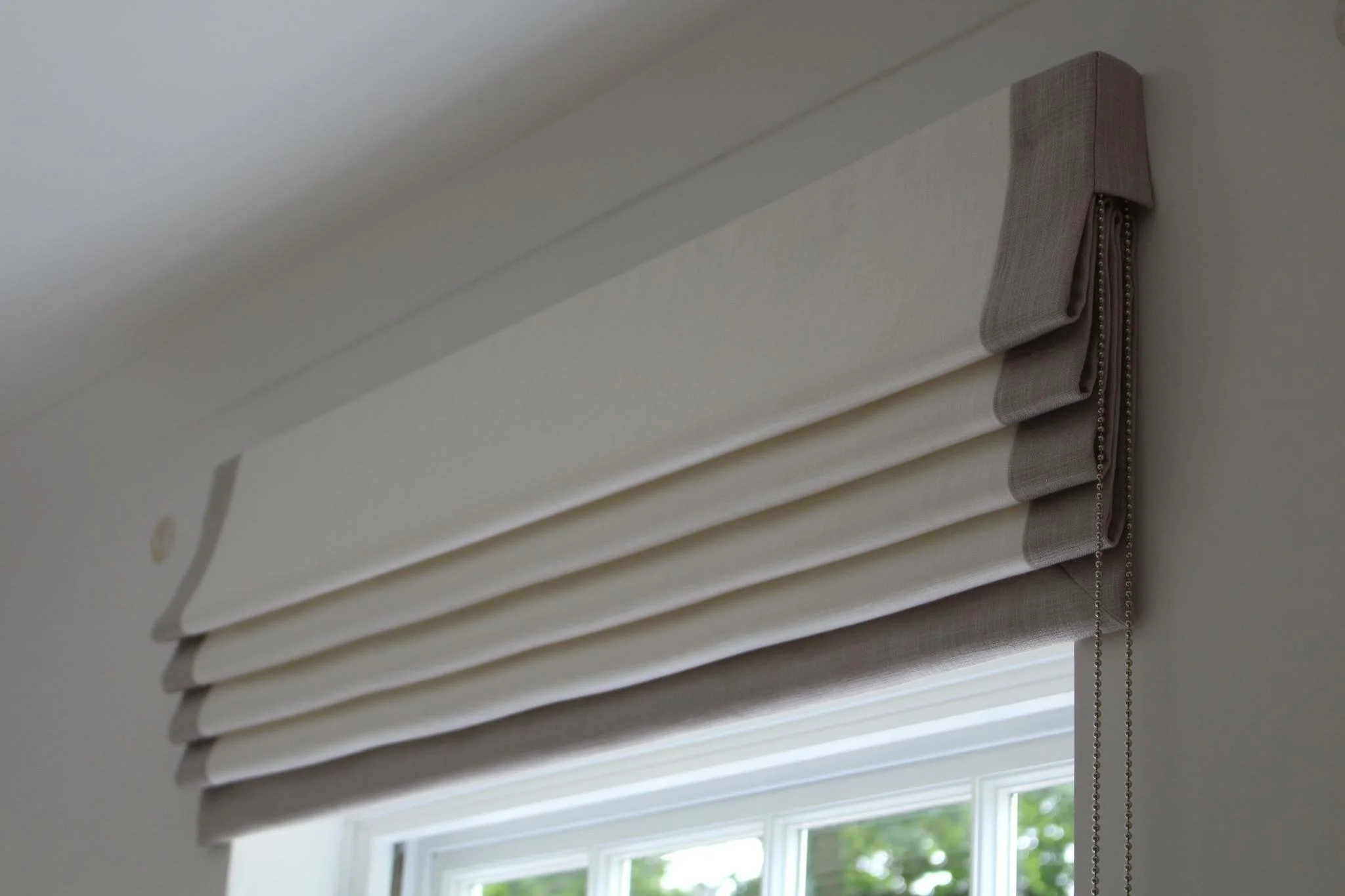 Close-up of a beige fabric window valance with side chains hanging down, mounted above a window with white trim, allowing natural light to enter.