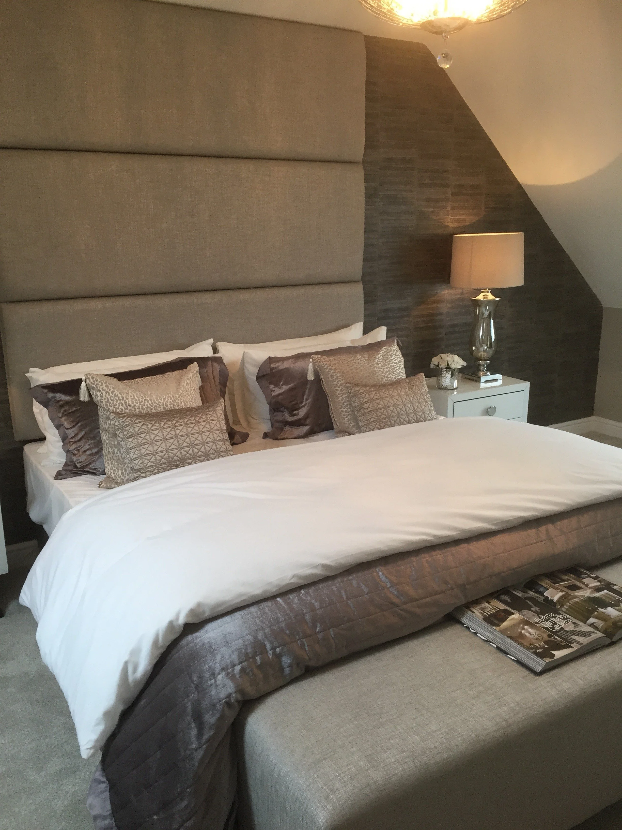 A luxurious bedroom with a large bed, multiple decorative pillows, a padded headboard, two bedside tables, a lamp, and a ceiling light.