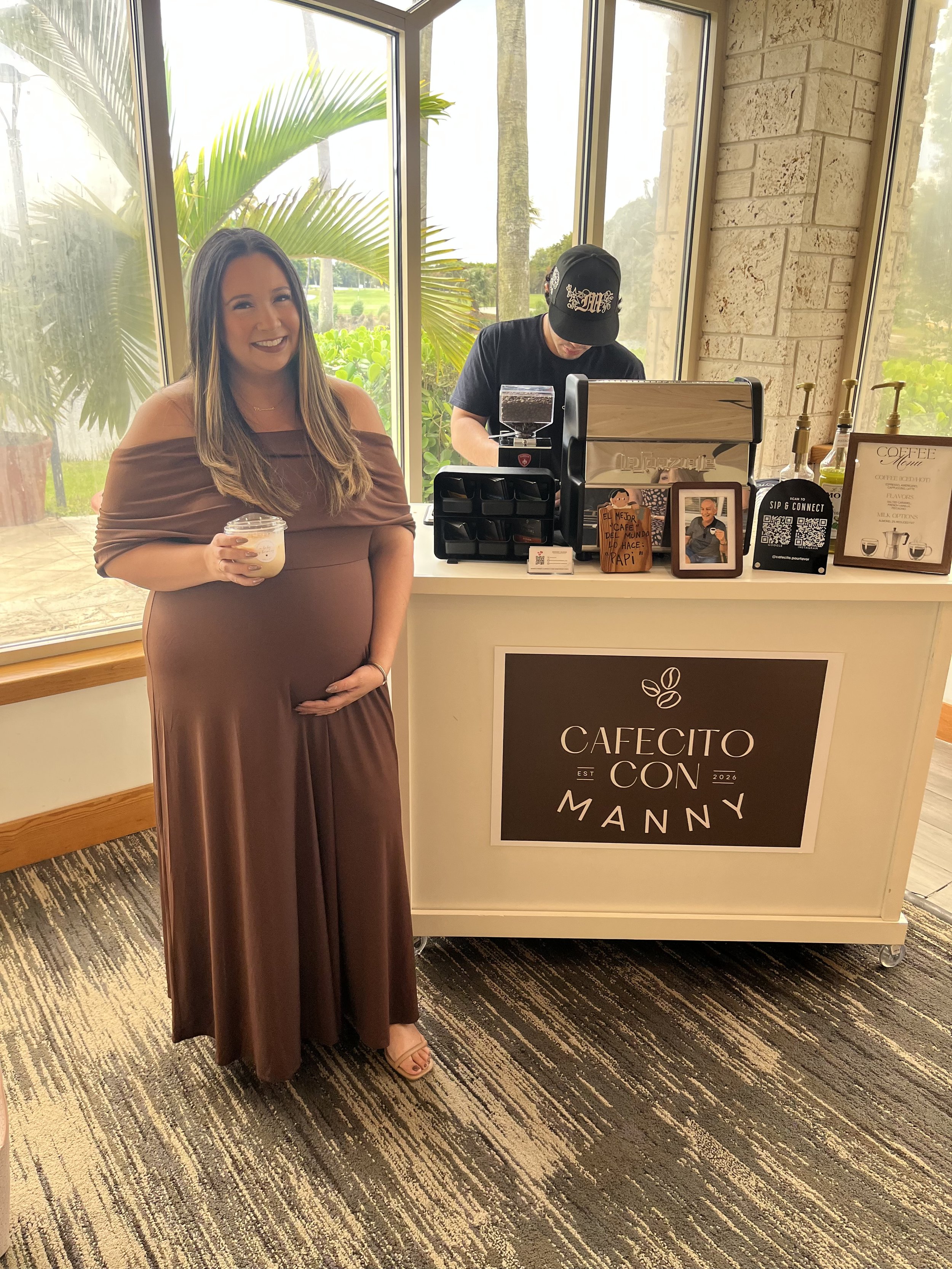 Pregnant guest smiling in front of a Cafecito Pour Favor coffee cart at a baby shower event in Miami