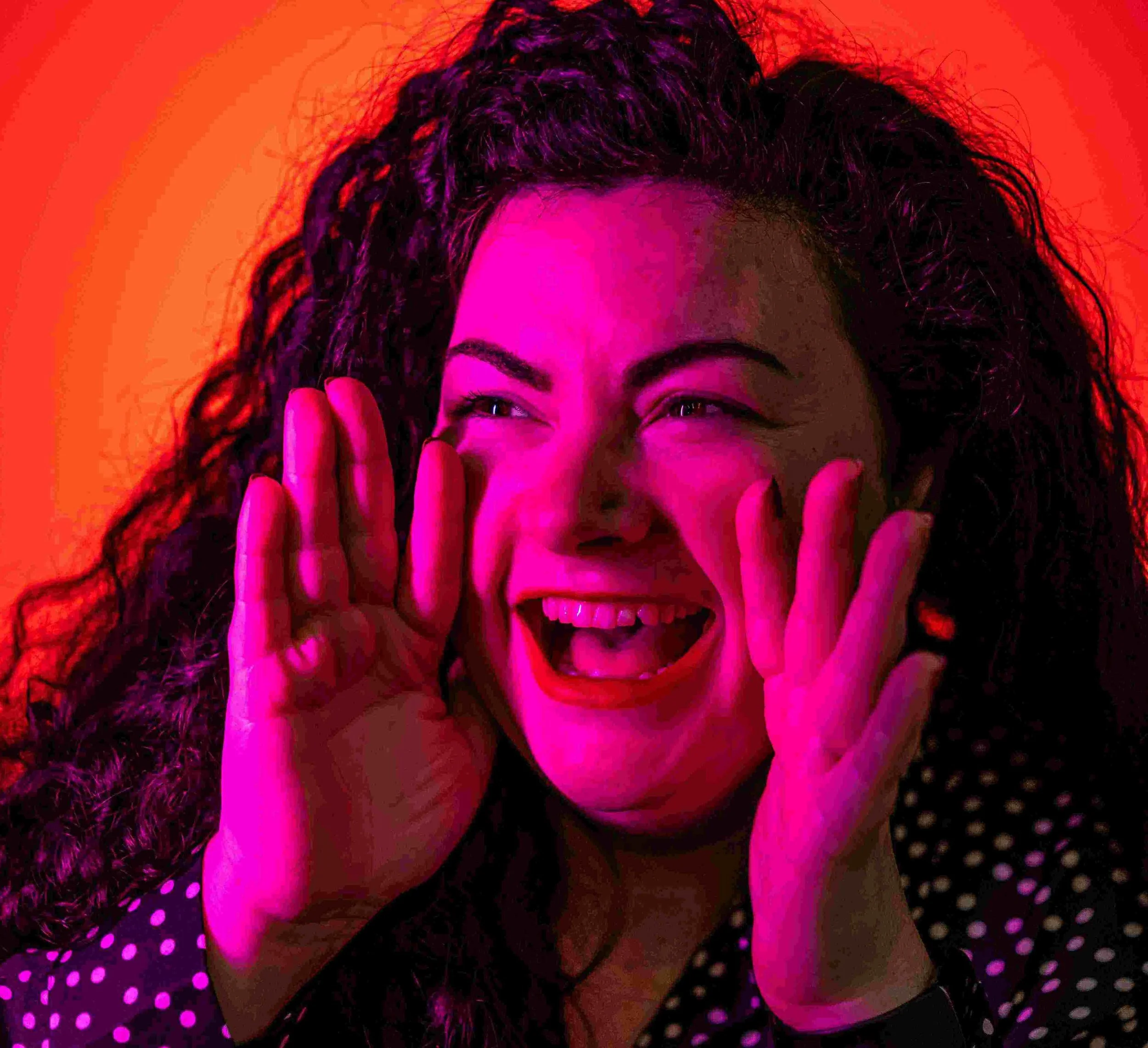 Marketing Meg, a woman with curly dark hair, smiling and laughing, holding her hands near her face against a vibrant pink and orange background, looking like she is shouting