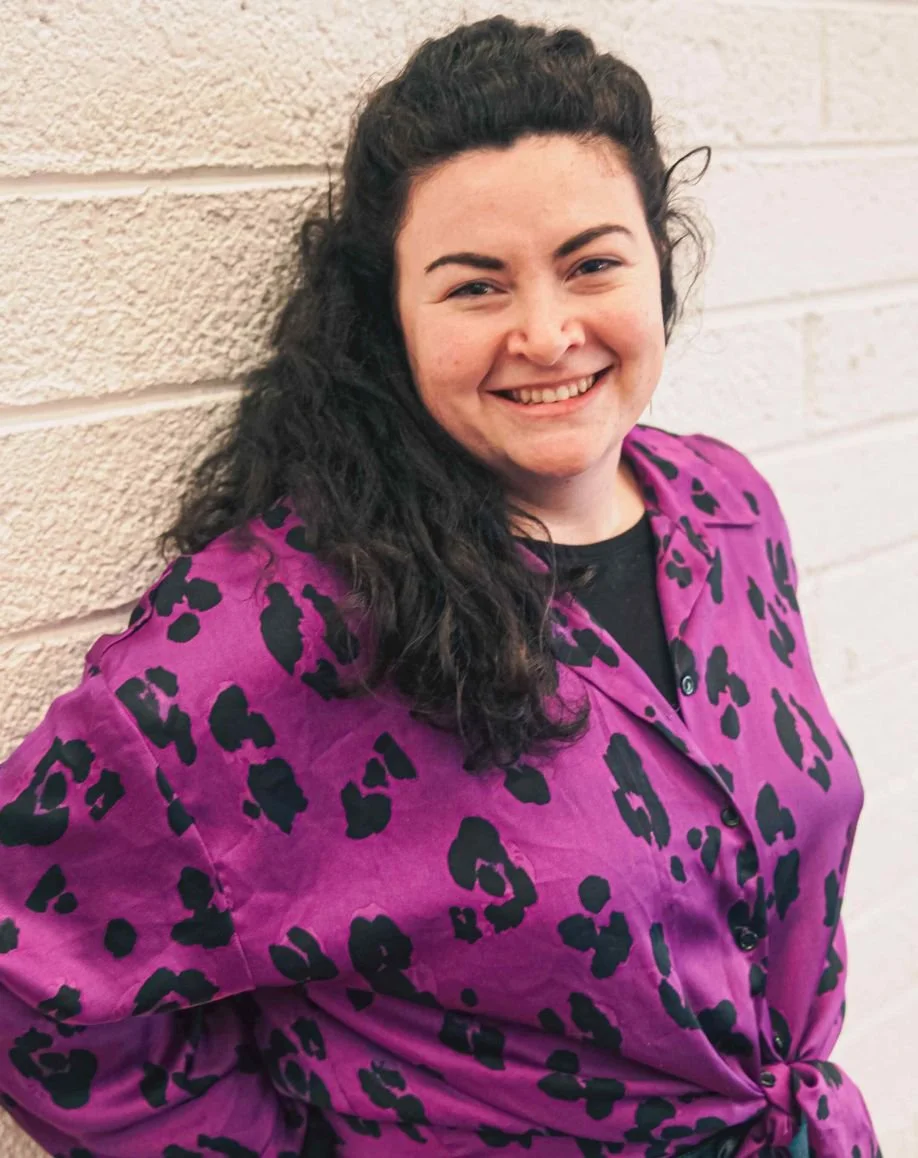 Marketing Meg, a woman with dark curly hair smiling and leaning against a beige brick wall, wearing a purple shirt with black leopard print.