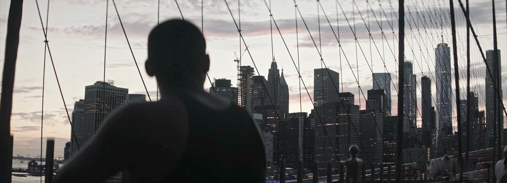 Silhouette of a person on a bridge with the New York City skyline in the background at sunset.