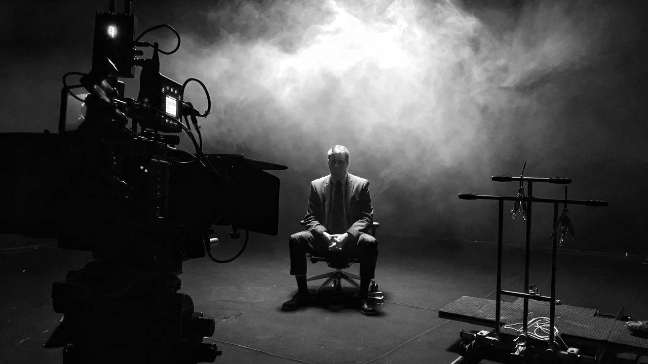 A man in a suit sitting on a chair on a dark stage, with stage equipment and a camera in front of him, and fog or smoke creating a moody atmosphere.