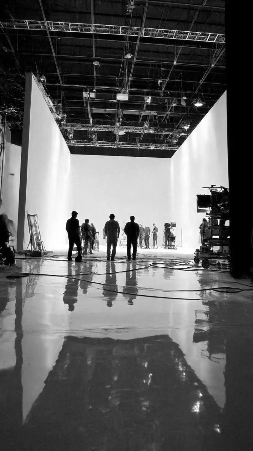 A behind-the-scenes view of a film or television studio with crew members working in front of a large white cyclorama wall, equipment and cables visible on the floor, and a high ceiling with lighting rigging, all in black and white.