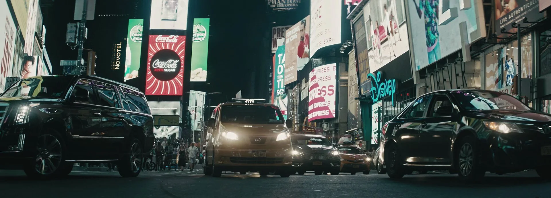 Nighttime street scene in Times Square, New York City, with cars and pedestrians. Bright digital billboards display advertisements, including Coca-Cola, Disney, and other brands.