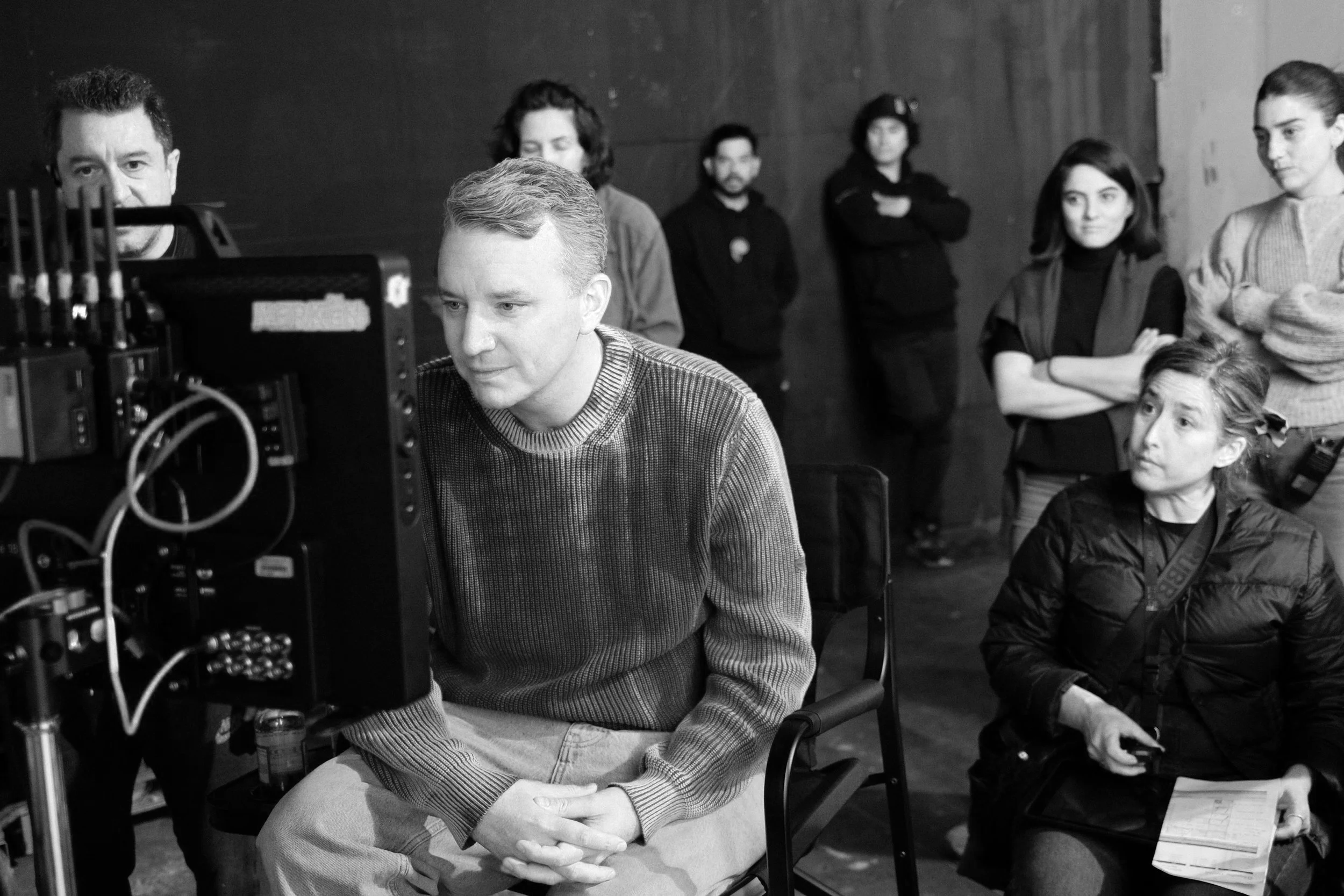 Black and white photo of a film or television production scene, with a man sitting and looking at a monitor, surrounded by crew members on a set or studio.