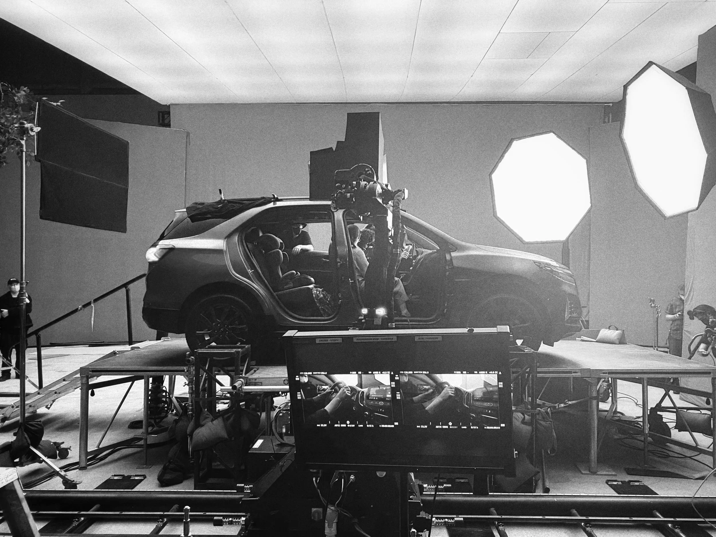 Black and white image of a car on a film set, surrounded by lighting equipment, cameras, and crew members. The car is positioned on a platform with crew members working around it.
