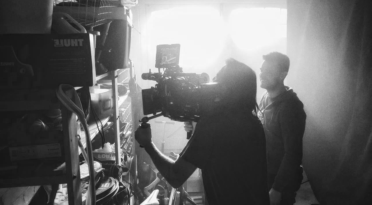 Film crew filming inside a storage room with shelves filled with boxes and equipment, backlit by bright light.
